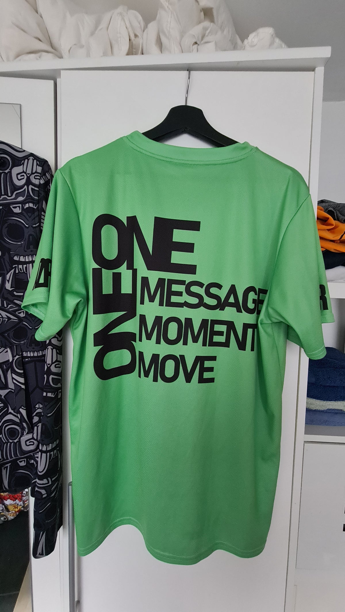 A bright green short-sleeve AZPR t-shirt hanging on a black hanger against a white closet door. The back of the shirt features large black text that reads 'ONE MESSAGE ONE MOMENT ONE MOVE'.